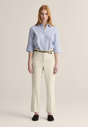 Kvinde med rødt hår iført en blå- og hvidstribet skjorte med knapper, beige bukser med guldknapper, flettet bælte og mørke loafers.