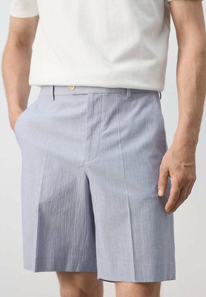 Hombre que lleva puestos unos pantalones cortos a medida de color azul claro con textura y una camisa blanca de manga corta, con una mano en el bolsillo y la otra al lado.