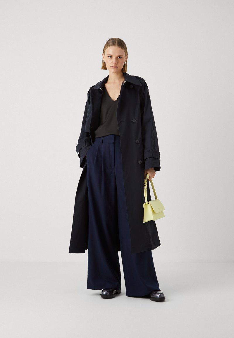 Lungo trench coat blu navy con una silhouette strutturata, abbinato a pantaloni a gamba larga e un top nero. Porta una piccola borsa gialla.