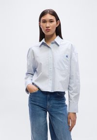 Ανοιχτό μπλε ριγωτό πουκάμισο με κουμπιά και τσέπη στο στήθος, με μακριά μανίκια και χαλαρή γραμμή, συνδυασμένο με μπλε τζιν παντελόνι.