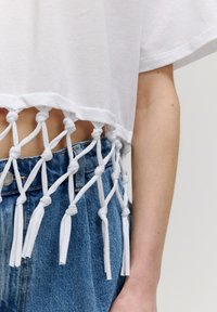 Witte cropped t-shirt met geknoopt stoffen detail aan de onderrand, gecombineerd met blauwe denim jeans. Stof lijkt zacht en heeft een casual ontwerp.