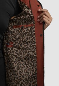 Interior de chaqueta marrón y roja con forro de estampado de leopardo. Presenta un bolsillo con un botón y un detalle de cremallera. Textura suave.