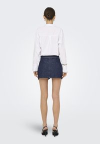 Chemise blanche à manches longues avec des poignets boutonnés, associée à une jupe courte en denim foncé. Le modèle porte des chaussures à talons hauts noires. Fond minimaliste.