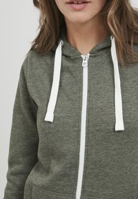 Sweat à capuche zippé vert olive en tissu doux, avec des cordons de serrage blancs, une poche avant et une fermeture éclair argentée. Design simple et décontracté.