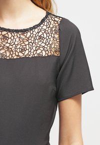 Svart kortärmad topp med en blommig spetsdesign över bröstet. Slätt, något glänsande tyg. Dekorativ halsringning med scallopkant.