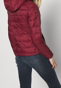 Czerwona kurtka puchowa z kapturem, z pikowanym wykończeniem i elastycznymi mankietami. Połączona z ciemnoniebieskimi dżinsami o gładkim wykończeniu.
