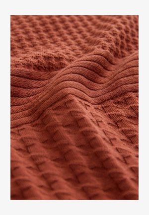 Tejido textil en color óxido con patrones texturizados. Presenta formas de diamante elevadas y líneas suaves y onduladas, mostrando un diseño de punto.