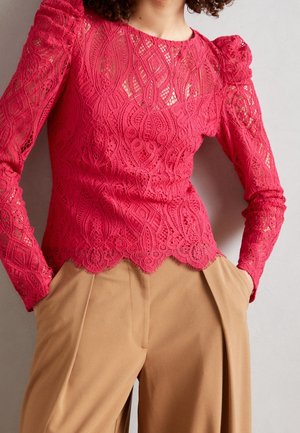 Vrouw draagt een felroze kanten top met lange mouwen en pofschouders en beige wijde broek, handen in zakken, tegen een effen achtergrond.