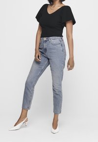 Haut noir côtelé avec manches courtes bouffantes, jean taille haute bleu clair, et escarpins slingback blancs à bout pointu. Accents de bijoux simples.