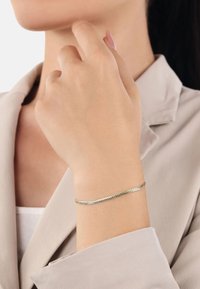 Tunn metallarmband med en tvinnad design, i guld- och silvertoner, bärs på handleden. Slät yta med en subtil glans.