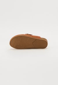Brown suede slip-on shoe with a textured rubber sole, featuring a curved shape and an adjustable strap for fit.