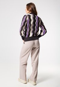Suéter de punto multicolor con patrones ondulados en púrpura, crema y negro; puños y cintura acanalados. Combinado con pantalones beige claro y zapatillas.