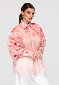 Femme aux cheveux foncés en queue de cheval basse, portant des boucles d’oreilles cœur dorées et un chemisier rose transparent à motifs floraux avec un pantalon blanc, debout devant un fond gris.