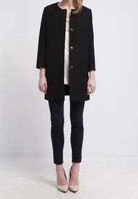 Abrigo negro con cuello redondo y tres botones, combinado con una blusa blanca y leggings oscuros, llevado con tacones nude.