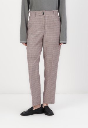 Personne portant un pantalon slim à carreaux avec boutons sur le devant, associé à des mocassins noirs et un haut gris à manches longues, debout devant un fond blanc.