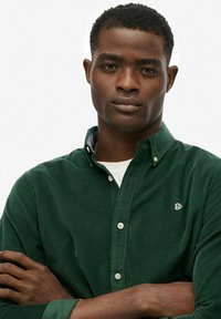 Joven con cabello corto y rizado, vistiendo una camisa de botones de color verde oscuro, de pie con los brazos cruzados frente a un fondo claro y liso.