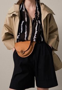 Tanfarvet lædertaske med rundet klap, båret over en sort plisseret nederdel, kombineret med en sort- og hvidstribet skjorte med knapper og en beige jakke.