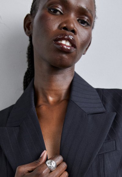 Zwarte gestreepte blazer met brede revers, met een diepe halslijn. Vingers versierd met een grote zilveren ring met textuur. Gladde huid en glanzende lippen.