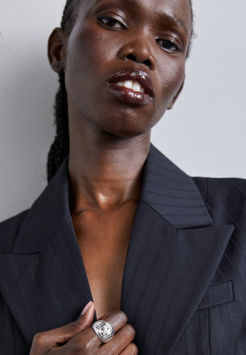 Fekete csíkos blézer széles hajtókákkal, mély dekoltázzsal. Az ujjakon egy nagy ezüst, textúrált gyűrű díszeleg. Simább bőr és fényes ajkak.