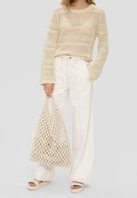 Pull beige en maille ajourée avec un motif à rayures horizontales, porté avec un jean blanc taille haute à jambes larges. Tenant un sac fourre-tout en tricot.