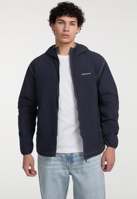 Giacca leggera navy con cappuccio, chiusura con zip, polsini elasticizzati e cuciture bianche. Abbinata a una maglietta bianca e jeans in denim sbiaditi.