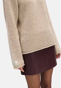 Beżowy sweter z dzianiny o luźnym kroju i długich rękawach, zestawiony z bordową skórzaną spódnicą. Etykieta widoczna na rękawie. Tkanina o fakturze.