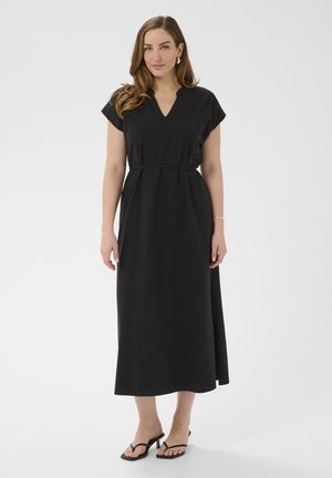 Naine seisab musta midi-kleit seljas, millel on lühikesed varrukad, V-kaelus, kitsas vöö ja mustad kontsakingad, taust on lihtne.