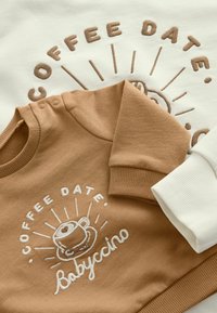 Brązowo-kremowe bluzy dziecięce z haftowanym kubkiem kawy i napisem „Coffee Date Babyccino” na przodzie, złożone ze skrzyżowanymi rękawami.