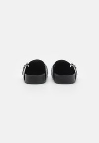 Schwarze Slipper mit strukturiertem Außenmaterial, offener Ferse und flacher Sohle. Dekorative Akzente an den Seiten verleihen dem Schuh Detail.