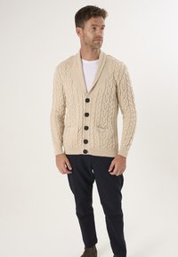 Beige strikket cardigan med kabelmønster, sjalskrave og seks sorte knapper. Har to frontlommer og ribbede manchetter.