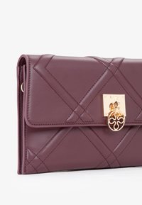 Portafoglio in pelle bordeaux con motivo trapuntato, chiusura a scatto in oro e un accento circolare. Presenta una chiusura a pattina con bordi cuciti.