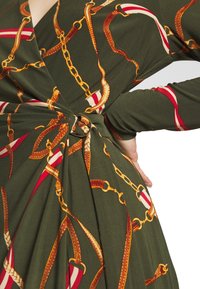 Wrapklänning i olivgrön tyg med guld kedjor och röda accenter. Texturerad yta med drapering och en cirkulär metalldetalj vid midjan.