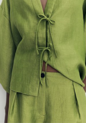 Haut en lin vert et pantalon assorti avec fermetures nouées, laissant apparaître un petit espace au niveau du ventre et un bouton noir sur la ceinture.
