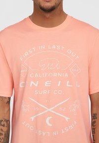Lys rosa bomulds t-shirt med et hvidt grafisk design med tekst, en bjørn, palmetræer, krydsede surfbrætter og et vintage-inspireret logo.