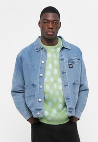 Denim jacket in light blue with silver buttons, breast pocket, and side pockets, layered over a green sweater with white polka dots.