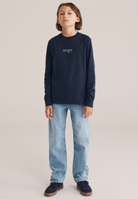 Námořnické tričko s dlouhým rukávem a bílou grafikou hory, kombinované s světle modrými džíny a tmavými botami. Jednoduchý design a uvolněný střih.