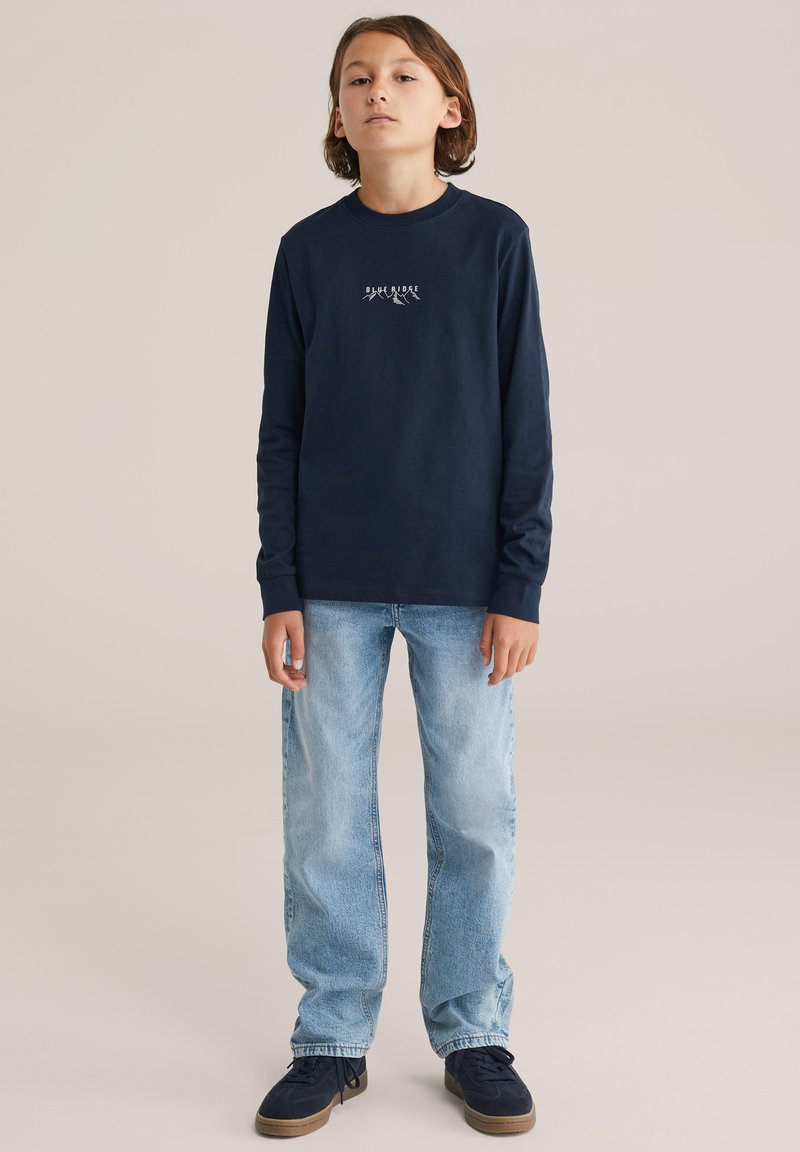 Sötétkék hosszú ujjú póló fehér hegy grafikával, világoskék farmernadrággal és sötét cipővel párosítva. Egyszerű dizájn és laza szabás.