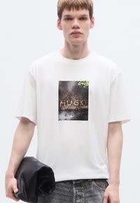 Λευκό βαμβακερό T-shirt με ένα τετράγωνο γραφικό σκούρου εδάφους με τη λέξη "HUGO" και πράσινες πινελιές. Το μοντέλο κρατάει μια μαύρη τσάντα χειρός.