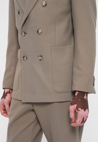 Khaki double-breasted blazer with textured fabric, large buttons, and pocket detail, paired with matching trousers and a hand with skin markings.