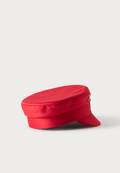 Casquette rouge à sommet plat avec une petite visière rigide et un petit détail de croix argentée sur le côté, sur un fond clair uni.