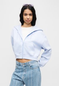 Νεαρή γυναίκα που φοράει μια ανοιχτό μπλε κοντή ζακέτα με φερμουάρ, ριγωτά ανοιχτό μπλε τζιν, λευκό τοπ και γυαλιά ηλίου στο κεφάλι, ποζάροντας μπροστά σε λευκό φόντο.