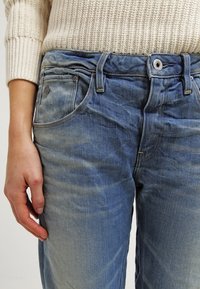 Jeans de mezclilla azul claro con un acabado desgastado, que cuentan con cinco bolsillos, cierre de botón y cremallera. La tela tiene una ligera textura.