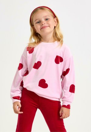 Roze fleece sweatshirt met grote rode hartmotieven, lange mouwen en een ronde halslijn, gecombineerd met rode strakke broek.