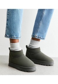 Botas de tobillo verde oliva hechas de un material suave, con punta redonda, puños acolchados y suelas texturizadas, combinadas con calcetines acanalados grises y jeans.