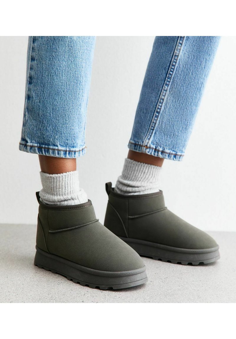 Botas de tobillo verde oliva hechas de un material suave, con punta redonda, puños acolchados y suelas texturizadas, combinadas con calcetines acanalados grises y jeans.