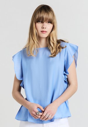 Blusa azzurro chiaro, con texture, maniche a volant e vestibilità ampia. Il tessuto presenta sottili righe verticali. Il modello indossa anelli d'argento sulle mani.
