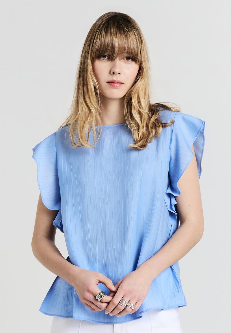 Blusa azzurro chiaro, con texture, maniche a volant e vestibilità ampia. Il tessuto presenta sottili righe verticali. Il modello indossa anelli d'argento sulle mani.