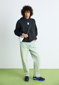 Young person with curly hair poses wearing a black hoodie, light jeans, black and blue sneakers, and carrying a small white crossbody bag.