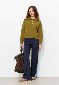 Pull olive avec un large col et trois boutons, associé à un pantalon évasé bleu marine avec des rayures latérales. Sac fourre-tout à carreaux marron.