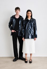 Twee personen dragen glanzende zwarte regenjacks met capuchons. De jacks hebben een gladde textuur en ritssluitingen. De complete outfits omvatten zwarte broeken en een witte rok.